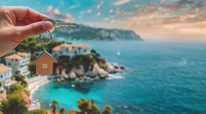 Hand holding house keys against a coastal village backdrop. New home ownership is highlighted with a stunning seaside view. Panorama with copy space..